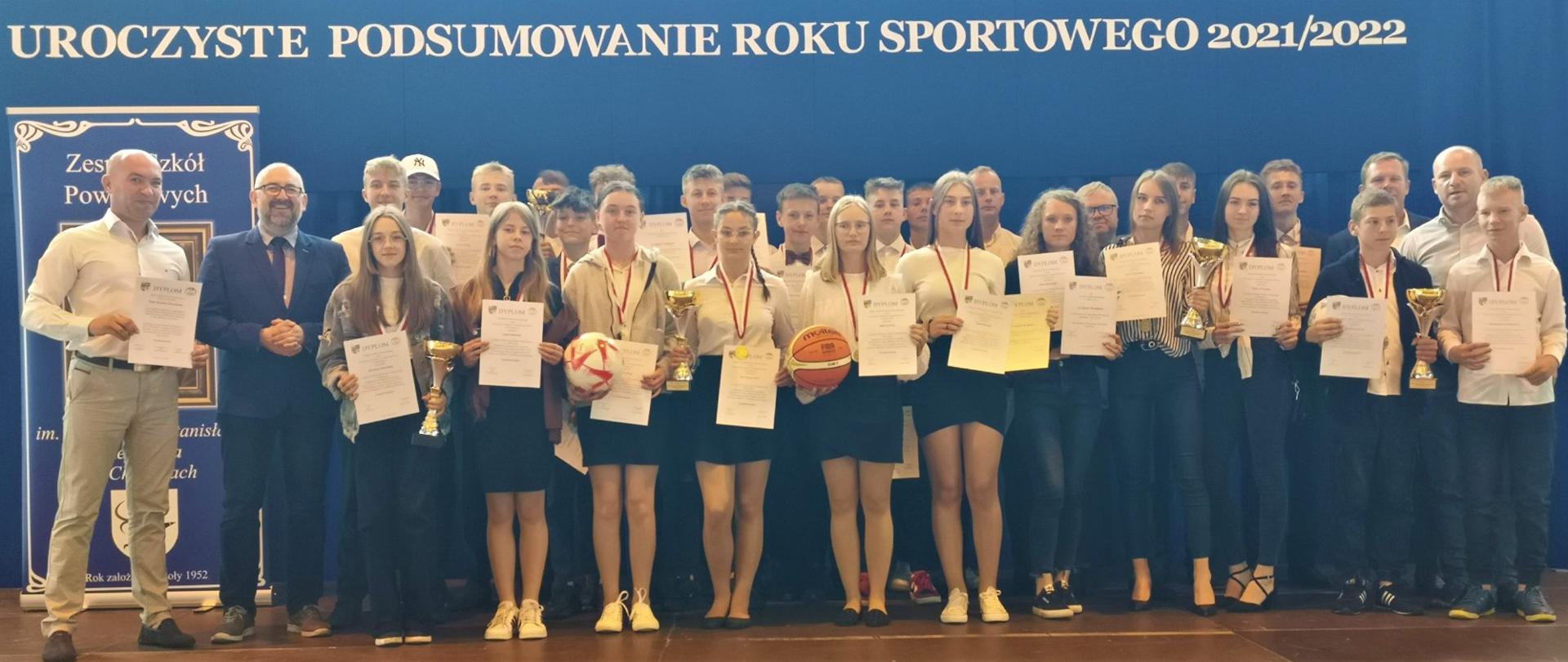 Na scenie stoją uczniowie z dyplomami, pucharami i odznaczeniami za zdobyte osiągnięcia sportowe. Wraz z nimi nauczyciele oraz Starosta Przasnyski Krzysztof Bieńkowski.