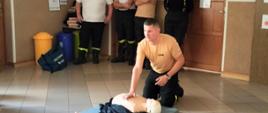 Na środku korytarza znajduje się mężczyzna ubrany w pomarańczową koszulkę z napisem: STRAŻ. Strażak nachyla się nad fantomem leżącym na niebieskim materacu. Jego twarz jest skierowana w stronę uczniów siedzących wokół niego na materacach.