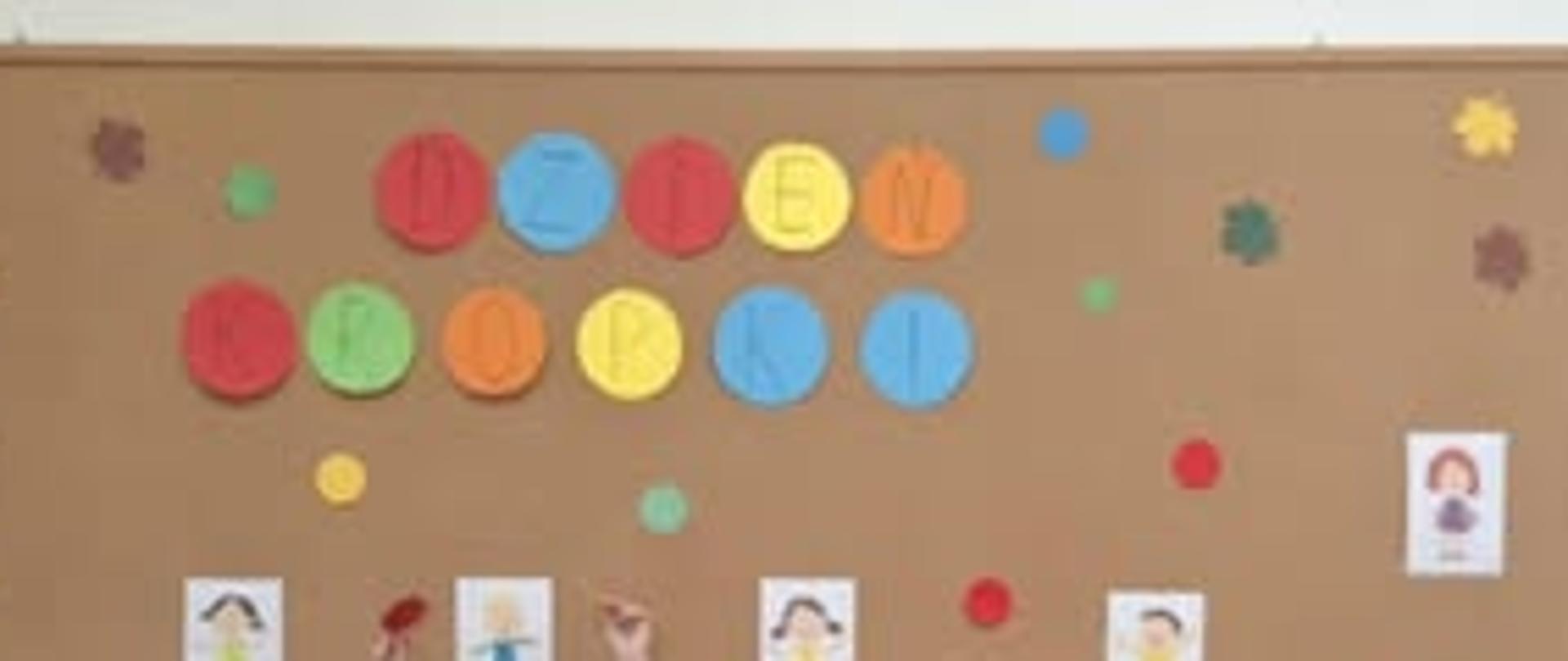 Grupka dzieci w dwóch rzędach ( jedni siedzą po turecku na ziemi a inni stoją za nimi) stoi pod tablicą korkową na której na kolorowych kropkach umieszczony jest napis "Dzień Kropki".