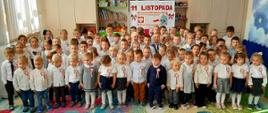 Duża grupa przedszkolaków stojąca razem w sali. Dzieci są ubrane w białe koszule i mają przypięte do ubrań biało-czerwone kotyliony lub kokardy, co sugeruje obchody Narodowego Święta Niepodległości. W tle widoczna jest dekoracja z napisem "11 LISTOPADA".