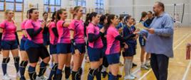 Fotografia na hali sportowej po lewej stronie drużyna w koszulkach różowych stoi w kolejce przed przewodniczącym Rady Powiatu Sokołowskiego, który składa gratulacje i wręcza dyplom i medale. 