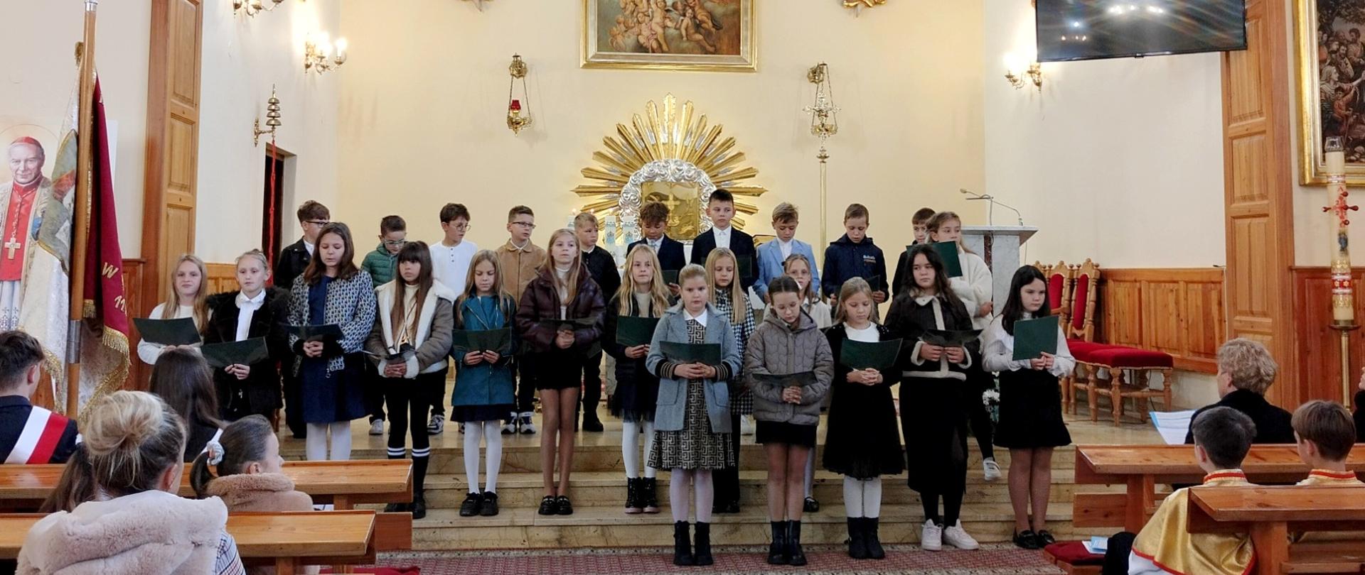 Uczniowie klasy czwartej stoją w kościele na schodach tyłem do ołtarza, ubrani w stroje galowe, prezentują program artystyczny z okazji rocznicy nadania szkole imienia
