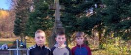 Trzech uczniów klasy 6 stoi przed ogrodzonym starym grobem z kamiennym krzyżem