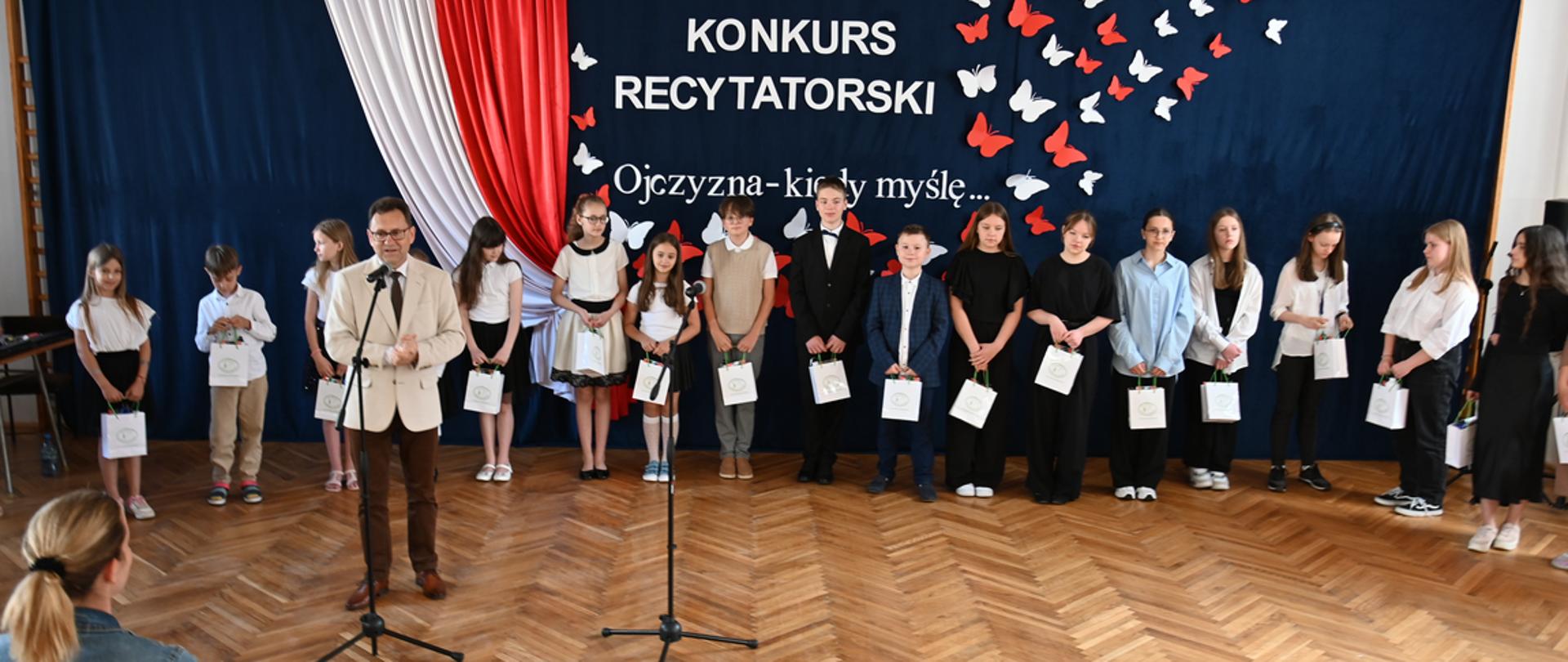 Na fotografii widzimy przemawiającego starostę, a za nim ustawionych w szeregu uczestników konkursu. W tle ścianka z biało - czerwoną flagą o oraz napis - tytuł konkursu.