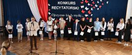 Na fotografii widzimy przemawiającego starostę, a za nim ustawionych w szeregu uczestników konkursu. W tle ścianka z biało - czerwoną flagą o oraz napis - tytuł konkursu.