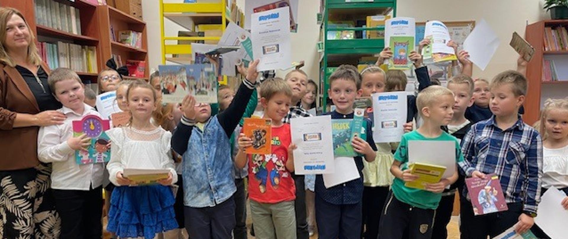 Grupa dzieci stojących w bibliotece szkolnej razem z nauczycielką. Dzieci trzymają w górze książki i dyplomy, wyrażając radość i świętując. W tle widać regały pełne książek.