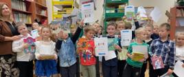Grupa dzieci stojących w bibliotece szkolnej razem z nauczycielką. Dzieci trzymają w górze książki i dyplomy, wyrażając radość i świętując. W tle widać regały pełne książek.