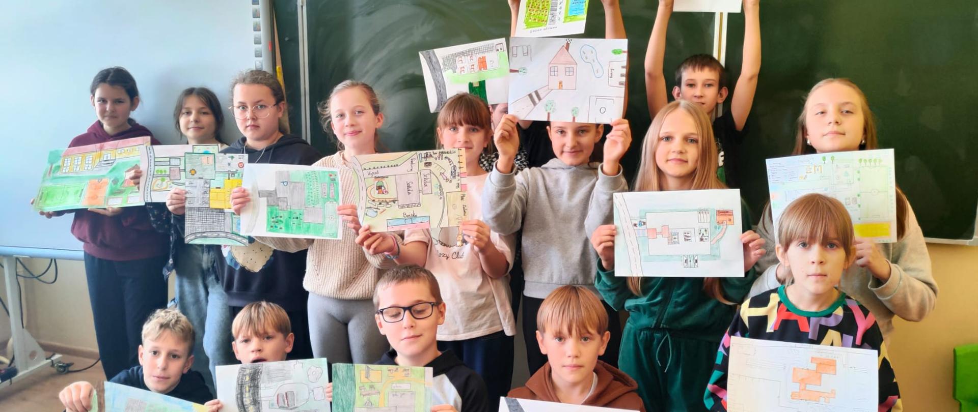 Uczniowie stoją w klasie, trzymają w ręku plakaty przedstawiające plany zagospodarowania terenu wokół szkoły, w tle tablica szkolna 