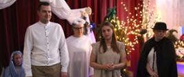 Wychowankowie ( 5 osób) w strojach teatralnych stoją na scenie przystrojonej dekoracjami świetlnymi i elementami świątecznymi. W tle widoczne są czerwone kurtyny.