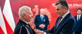 Na zdjęciu Wojewoda Lubelski (po prawej) przypina odznaczenie Staroście Kraśnickiemu. Starosta ubrany jest w galowy mundur strażacki. Na drugim planie znajdują się biało-czerwone flagi oraz czerwona ścianka Państwowej Straży Pożarnej. Na ściance znajduje się biały orzeł w koronie.