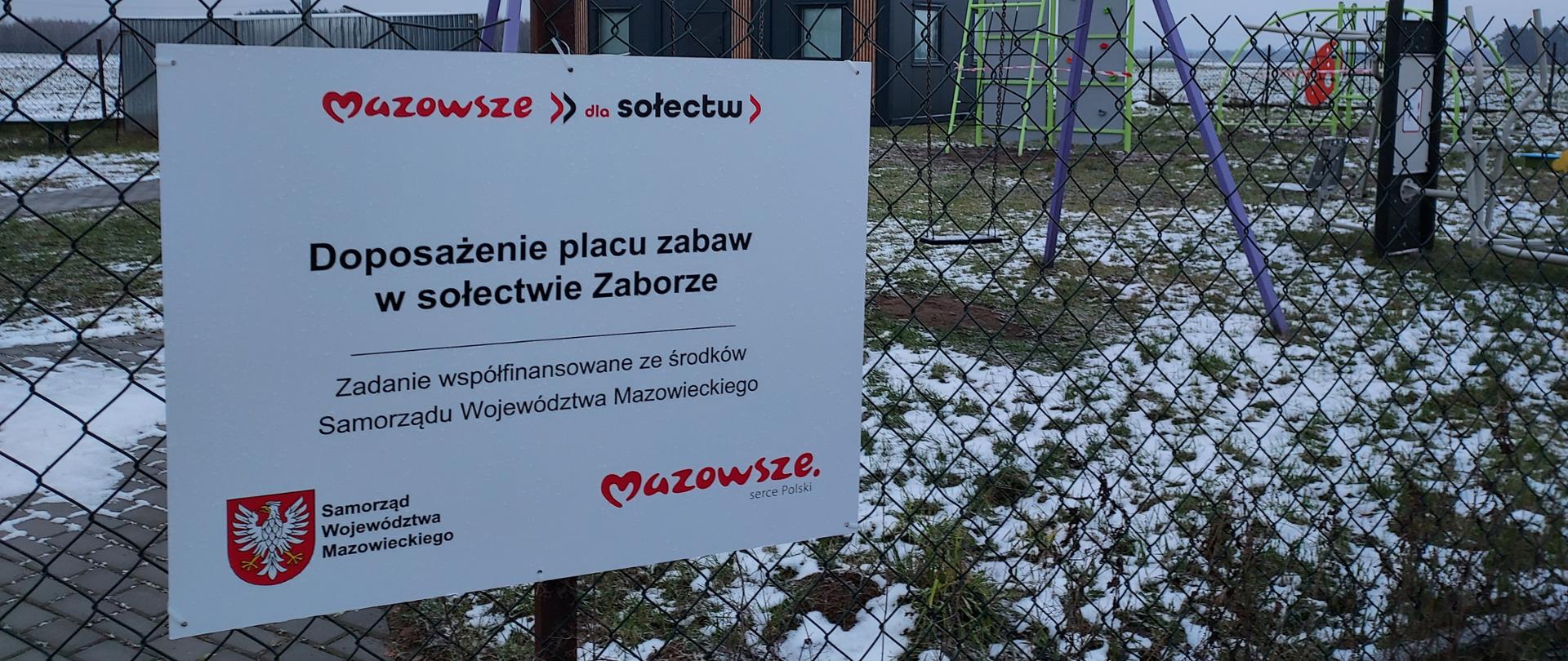 Zdjęcie przedstawia tablicę informacyjną o treści: Doposażenie placu zabaw w sołectwie ZAborze - zadanie współfinansowane ze środków Samorządu Województwa Mazowieckiego. Na zdjęciu umieszczono logo marki Mazowsze oraz logo programu "Mazowsze dla sołectw". W dali widoczne zamontowane urządzenia na placu zabaw.