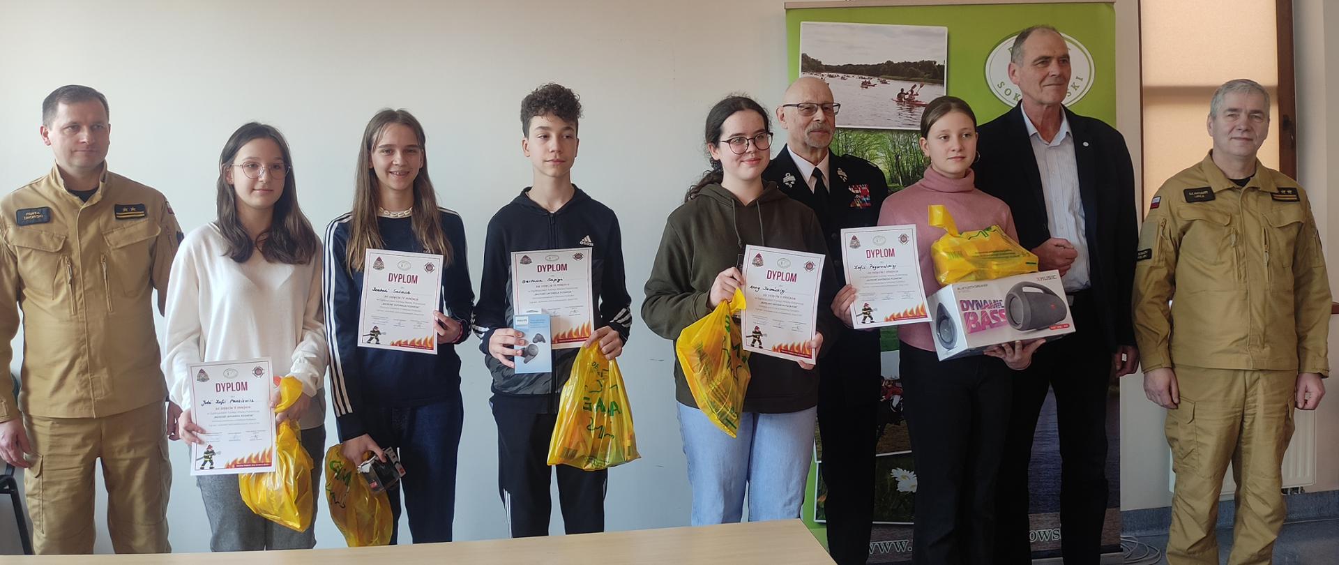 Zdjęcie grupowe laureatów turnieju wraz z członkami Jury na tle rollupu z logo Powiatu Sokołowskiego. W pierwszym rzędzie piątka dzieci z dyplomami
w dłoniach oraz z reklamówkami z nagrodami. Z tyłu członkowie komisji. Dodatkowo na zdjęciu w drugim rzędzie wicestarosta Ryszard Domański, który wręczał młodzieży nagrody.
