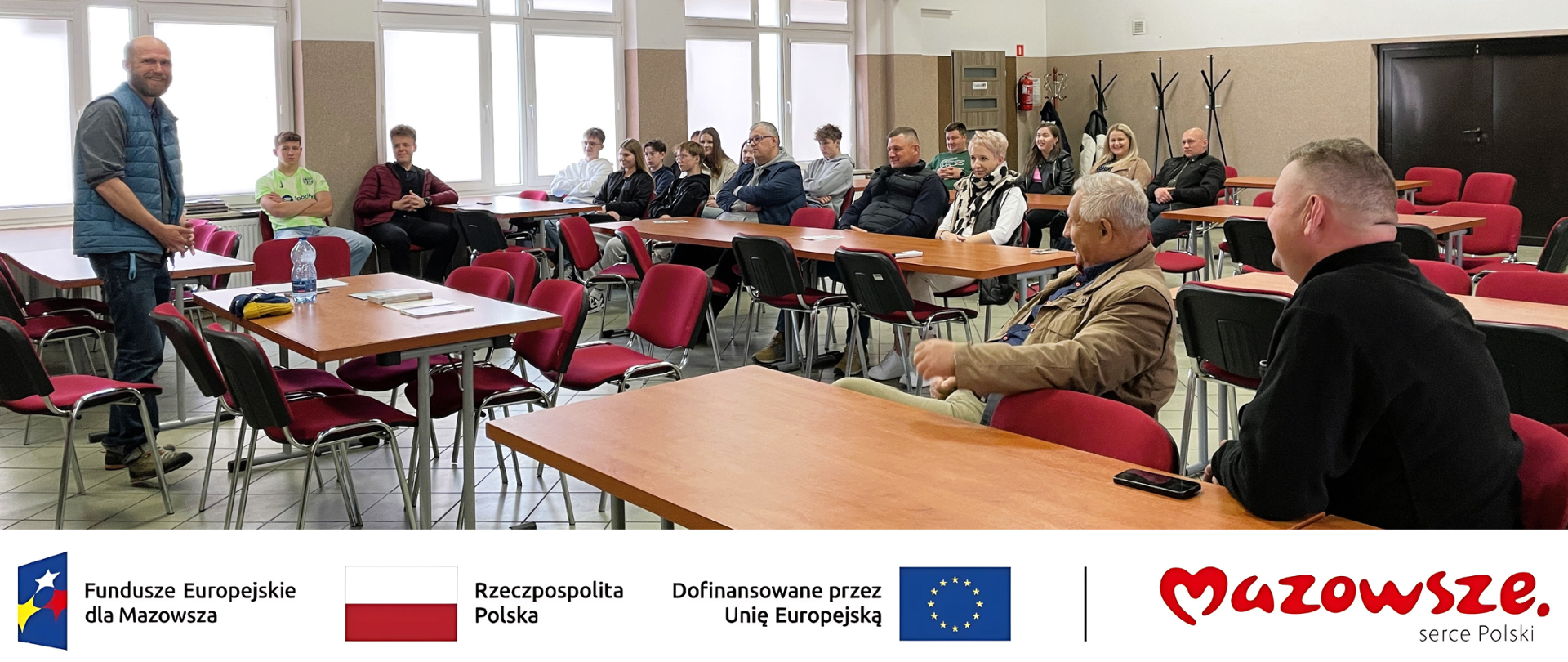 Wnętrze sali osp, mężczyzna stoi przed zgromadzonymi uczestnikami spotkania siedzącymi przy stolikach. Na dole zdjęcia znajdują się logotypy: Funduszy Europejskich dla Mazowsza, Flaga Polski, informacja o dofinansowaniu przez Unię Europejską z flagą UE i logotyp Mazowsze serce Polski.