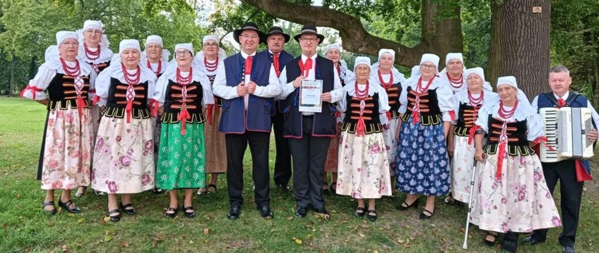 Zespół w śląskich strojach pozuje do zdjęcia na polanie pod drzewem.