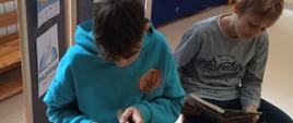 Dwóch uczniów z klasy 8 czyta na przerwie podczas akcji bibliotecznej "Przerwa na czytanie", w tle gazetka promująca wydarzenie