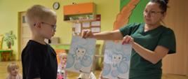Na zdjęciu widać opiekunkę i pięcioro dzieci z grupy Tygryski. Opiekunka trzyma w ręku dwie ilustracje. Jedna przedstawia uśmiechnięty ząbek a druga smutny.