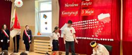 Uczeń i uczennica podchodzą do ucznia przebranego za króla w złotej koronie na głowie, który siedzi na tronie wyraźnie zasmucony, po lewej stronie poczet sztandarowy ze sztandarem szkoły