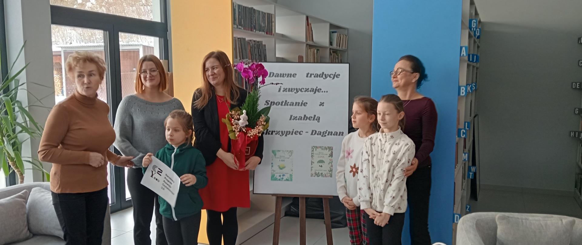 Od lewej: bibliotekarka ze szkoły w Nowej Wsi, pani dyrektor Gminnej Biblioteki Publicznej w Łabowej, obok pisarka stojąca przy stojaku ze swoim nazwiskiem, po prawej stronie bibliotekarka z biblioteki filialnej oraz trzy dziewczynki, które wręczały pisarce kwiaty