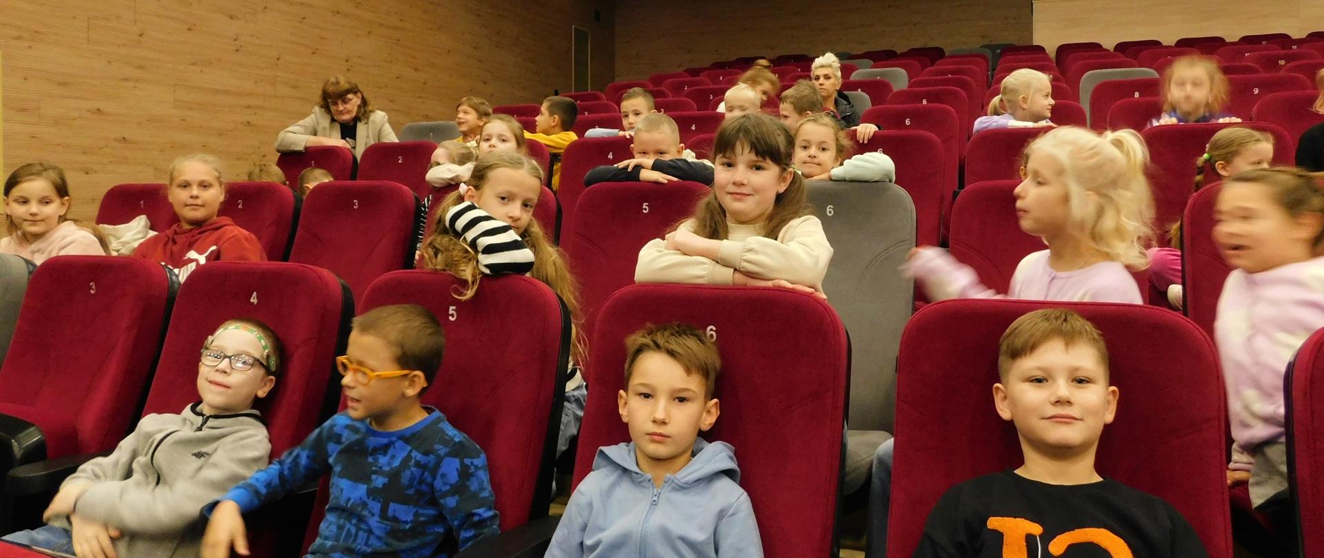 U dołu zdjęcia, na bordowych fotelach siedzą w rzędach dzieci. U góry zdjęcia widnieją lampy oświetlające salę teatru.