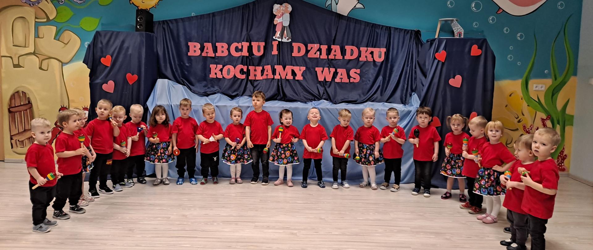 Dzieci z grupy Delfinki ustawione w półkolu, na tle granatowej dekoracji dla Babci i Dziadka, ubrane w czerwone koszulki. W ręku trzymają instrumenty muzyczne. 
