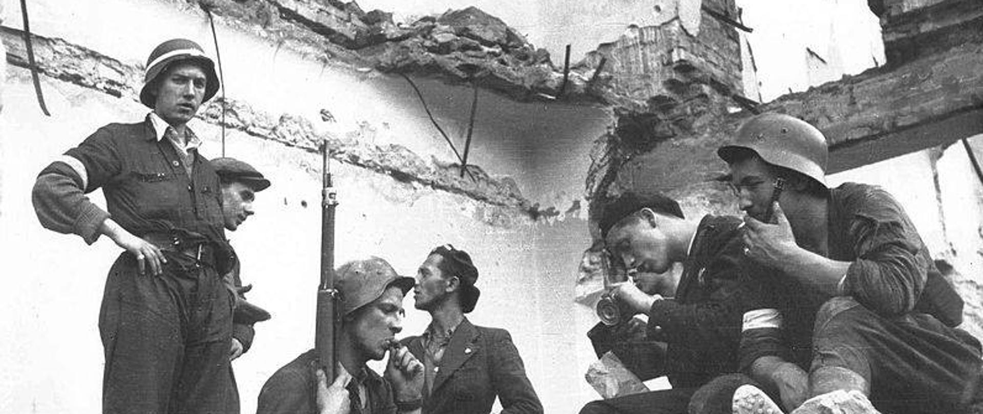 Zdjęcie przedstawia sześć osób w mundurach siedzi na gruzach budynku. Zdjęcie czarno-białe przedstawia Powstańców Warszawskich