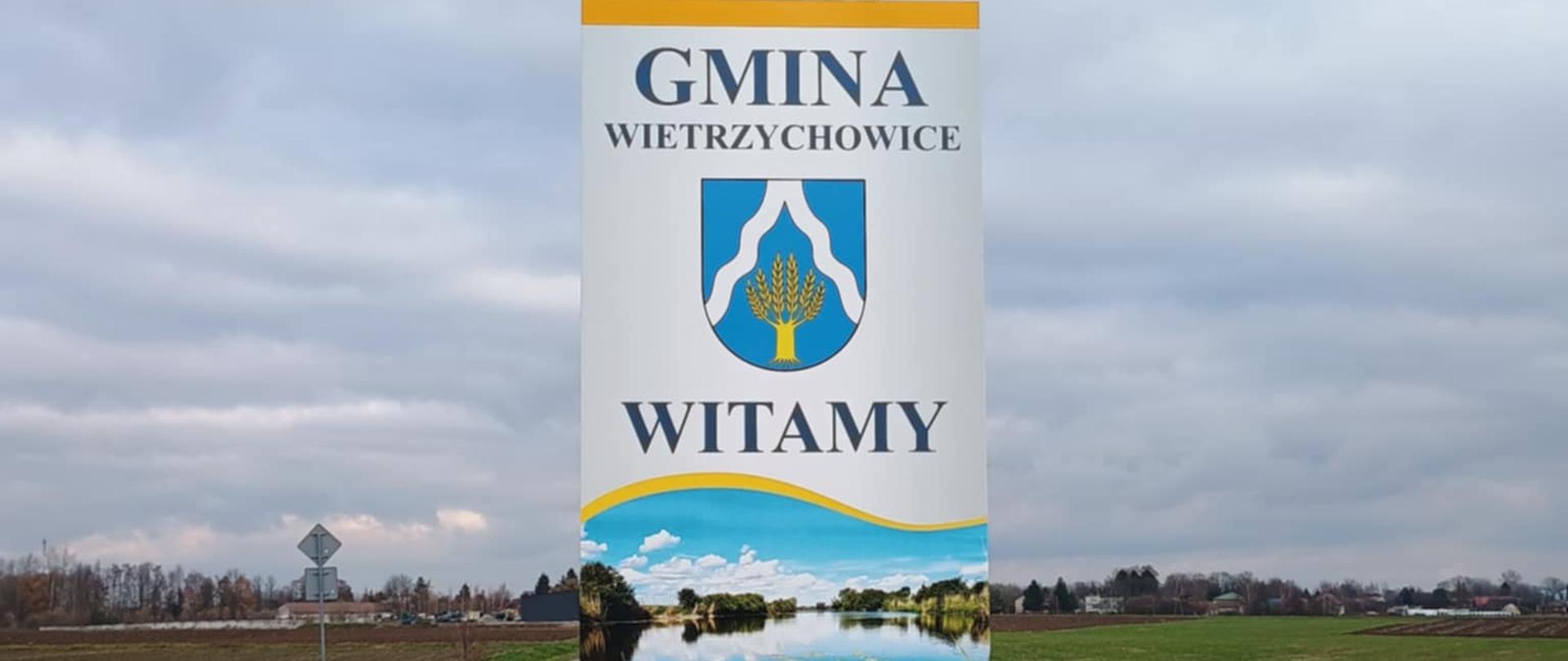 Zdjęcie przedstawia na dwóch metalowych rurkach tablicę informacyjną, na której znajduje się napis Gmina Wietrzychowice, herb, napis Witamy oraz zdjęcie charakterystycznego miejsca w gminie, czyli Jeziora Święconego. W tle znajdują się zabudowania gospodarstw domowych.