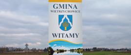 Zdjęcie przedstawia na dwóch metalowych rurkach tablicę informacyjną, na której znajduje się napis Gmina Wietrzychowice, herb, napis Witamy oraz zdjęcie charakterystycznego miejsca w gminie, czyli Jeziora Święconego. W tle znajdują się zabudowania gospodarstw domowych.