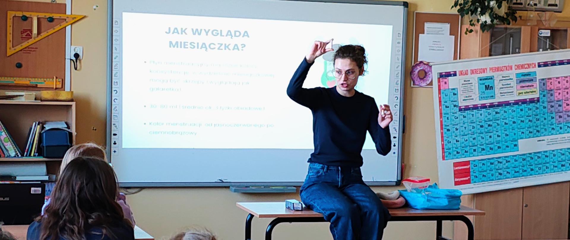 Kobieta siedzi na ławce szkolnej i ma uniesioną prawą rękę. Przed nią siedzą uczennice. Za nią tablica z napisem Jak wygląda miesiączka.