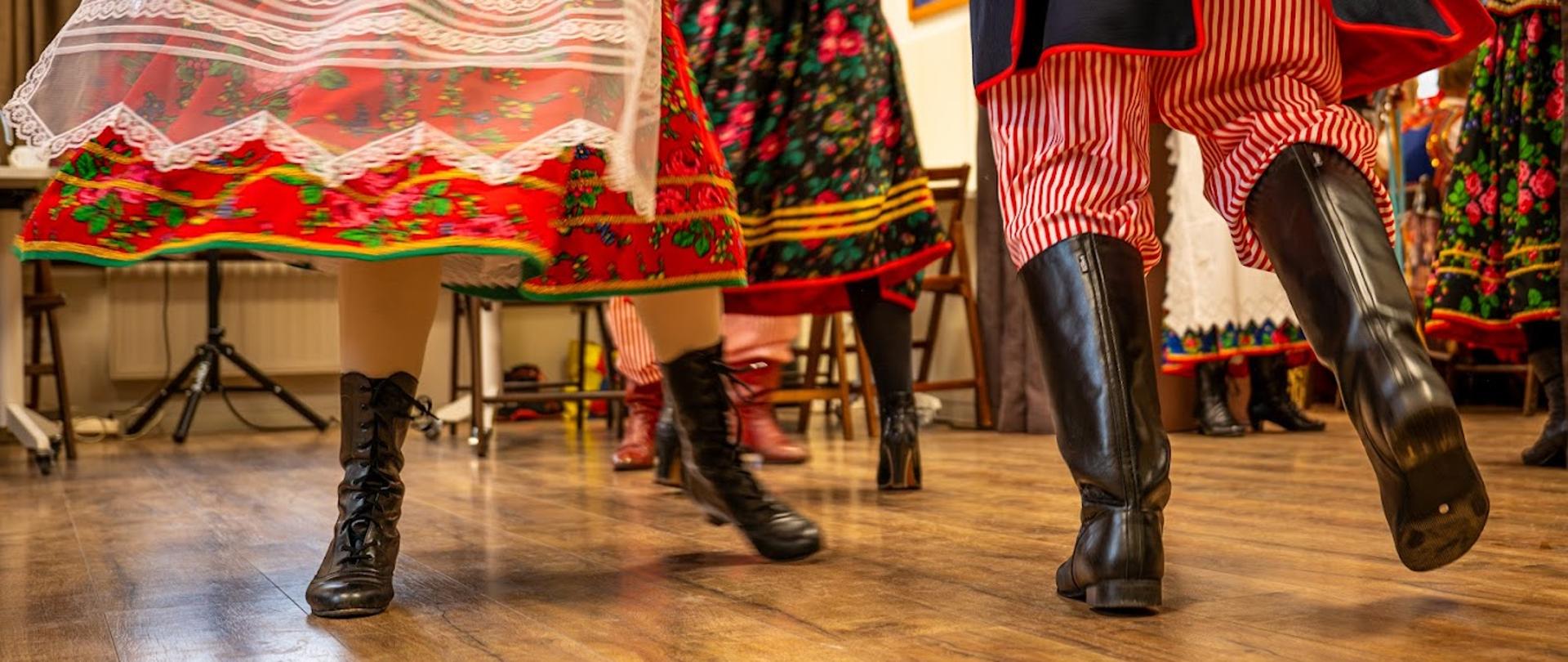 Tancerze wykonujący tradycyjny taniec ludowy w kolorowych strojach folklorystycznych i skórzanych butach na drewnianej sali.
