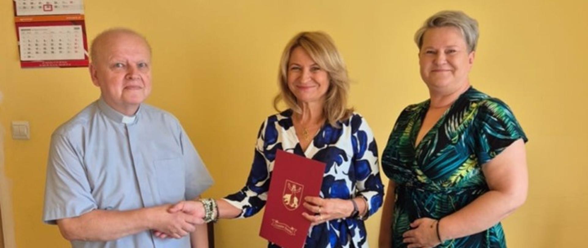 2 kobiety i ksiądz, uścisk dłoni, teczki z herbem gminy sawin