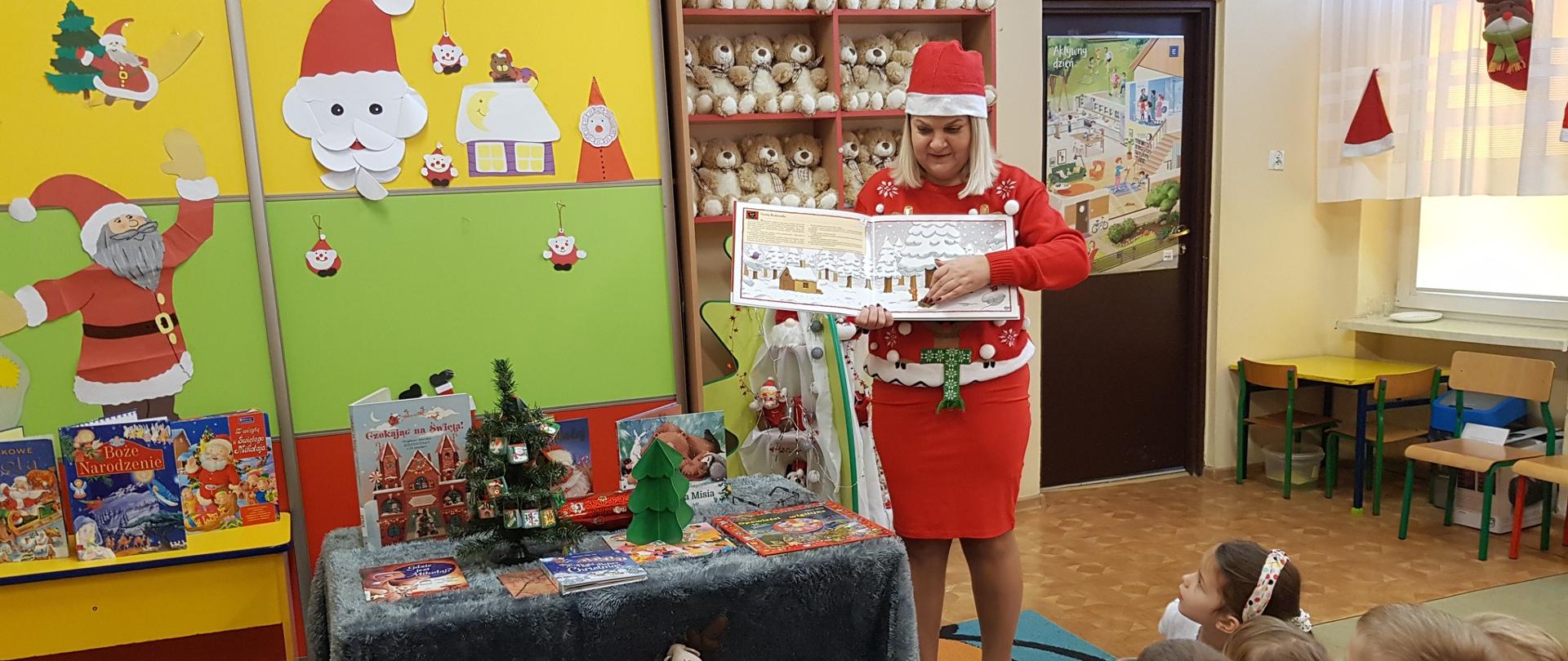 Pani w czerwonym stroju z czapką mikołajkową na głowie przedstawia dzieciom książeczkę z ilustracjami