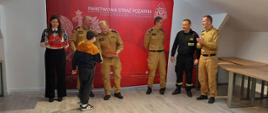 5 strażaków oraz kobieta. Przed nimi stoi chłopie i podaje rękę strażakowi który w drugim ręku trzyma czerwoną książkę