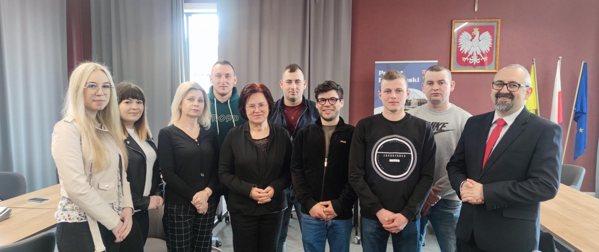 Zdjęcie przedstawia Starostę Przasnyskiego Krzysztofa Bieńkowskiego, Dyrektor Powiatowego Urzędu Pracy Jadwigę Żuber oraz Panią Barbarę Garlińską wraz z nowymi przedsiębiorcami powiatu przasnyskiego tuż po podpisaniu umów na dofinansowanie rozpoczęcia ich działalności gospodarczej. Zdjęcie wykonano na sali konferencyjnej Centrum Aktywizacji Przedsiębiorczości w Starostwie Powiatowym w Przasnyszu, w tle szare kotary, ściany w kolorze szlachetnego mahoniu oraz oprawione godło Rzeczypospolitej Polskiej – biały orzeł w złotej koronie na czerwonym tle.
