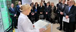 Zdjęcie przedstawia jedną osobę przemawiającą przy mównicy wewnątrz budynku. Przed nią stoi kilkadziesiąt osób