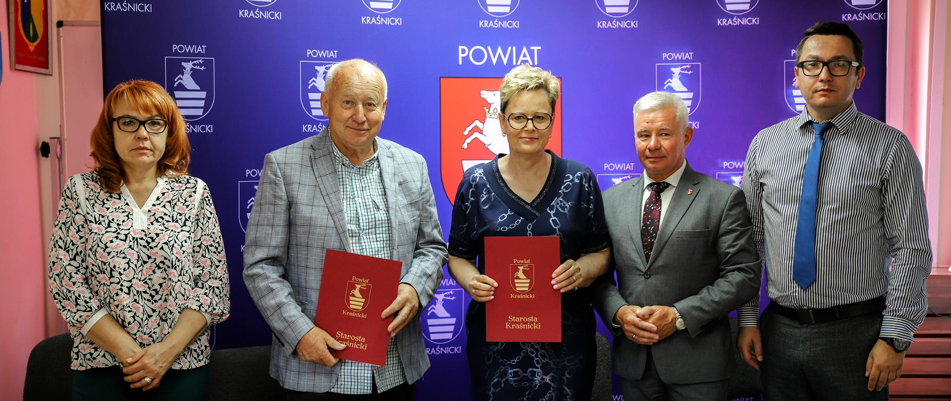 Zdjęcie przedstawia moment przekazania podpisanych umów na realizację zadania inwestycyjnego. Za stołem prezydialnym stoi pięć osób.