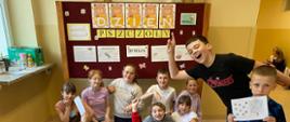 Uczniowie siedzą na podłodze pod gazetką tematyczną na stojaku. Na gazetce widoczny napis "Dzień Pszczoły" w formie liter trzymanych przez misie