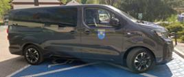 Boczny widok ciemnoszarego samochodu Opel Zafira przeznaczonego do transportu osób z niepełnosprawnościami. Pojazd posiada przyciemniane szyby oraz oznaczenie herbu Gminy Siepraw na przednich drzwiach. Samochód stoi na parkingu przed budynkiem użyteczności publicznej.