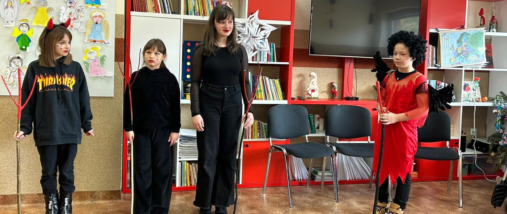 Na środku występujące w jasełkach 3 uczennice przebrane na czarno za diabły z czerwonymi rogami i widłami, po prawej uczeń przebrany za diabła na czerwono, w tle szafki z książkami, krzesła, telewizor