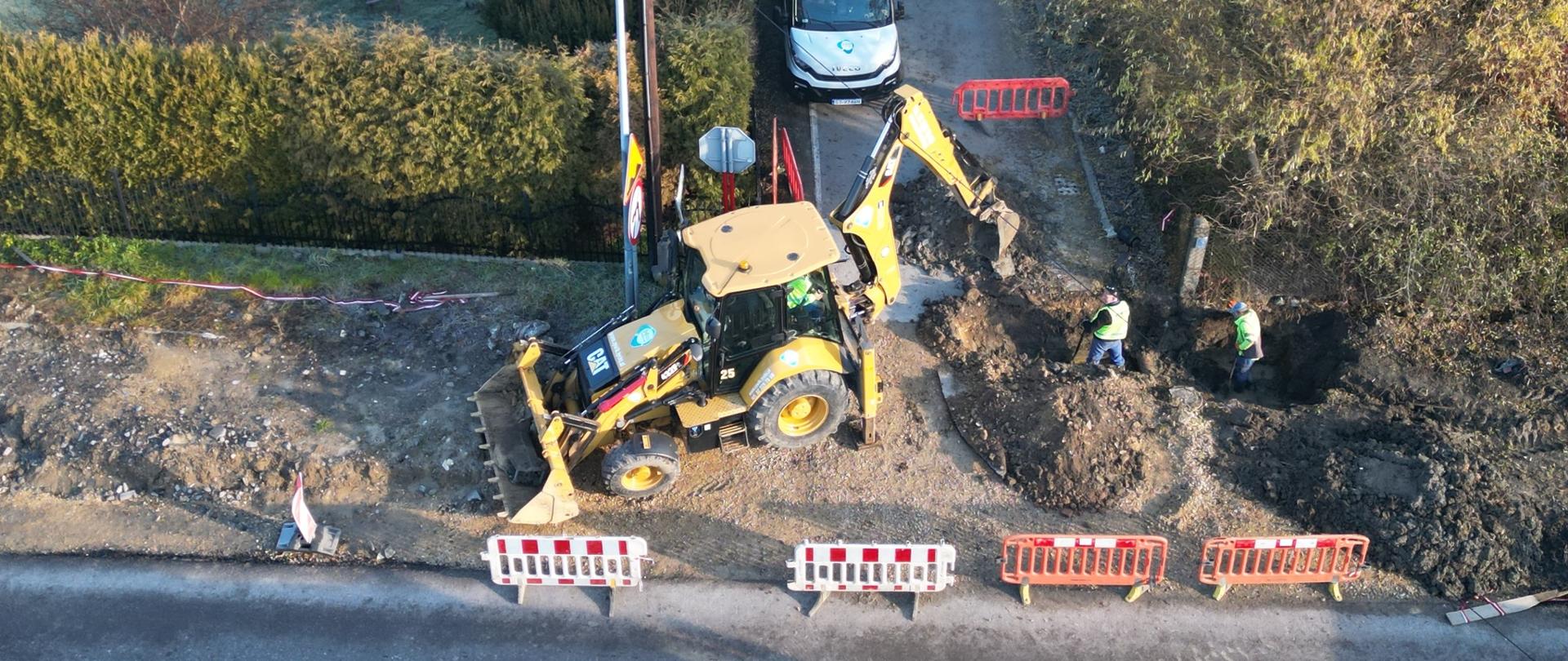 Koparka kopie dziurę w ziemi, robotnicy poprawiają wykop.