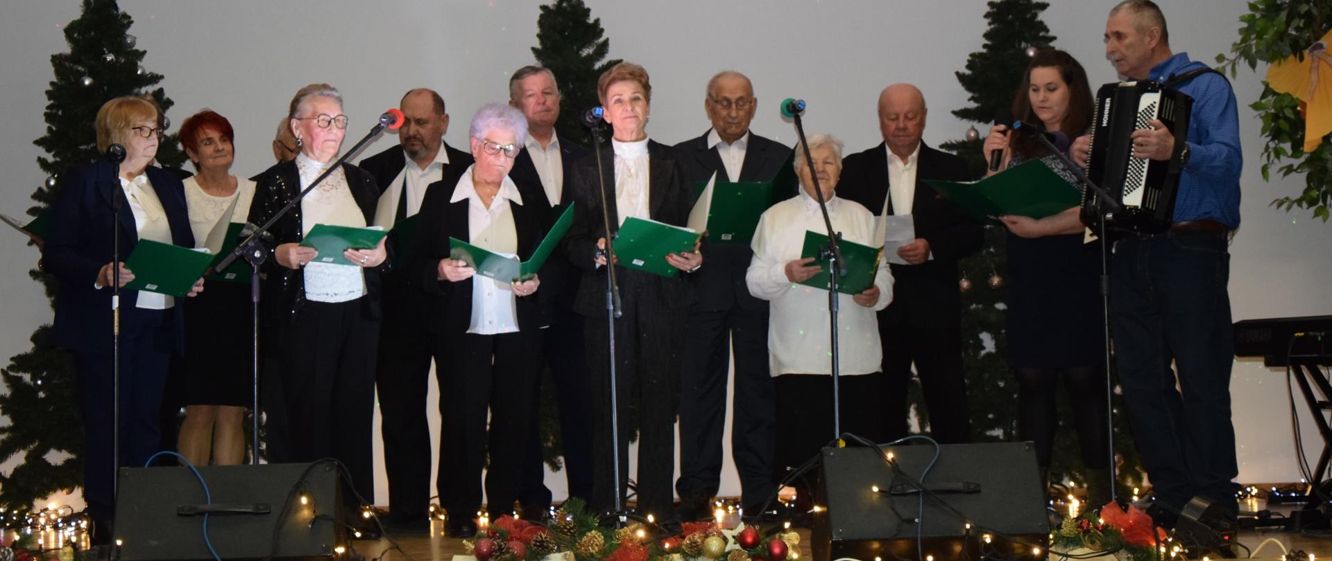 Zdjęcie przedstawia grupę seniorów (kobiet i mężczyzn) śpiewających na scenie.