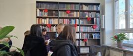 Uczniowie zwiedzają bibliotekę, za nimi potrójny regał z książkami, w tle nauczycielka przeglądająca książkę