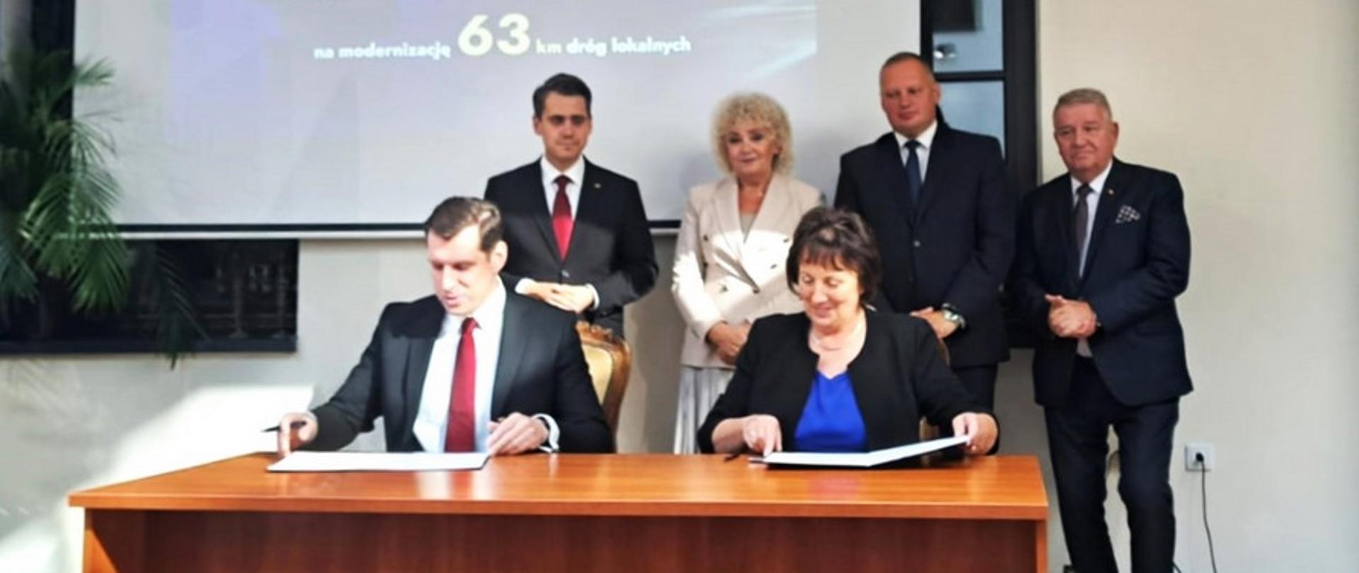 Na fotografii widzimy stolik, przy którym starosta Elżbieta Sadowska oraz Wojewoda Mazowiecki podpisują umowy. w tle stoją parlamentarzyści i widać ekran na którym wyświetlana jest informacja dotycząca Rządowego Funduszu Rozwoju Dróg.