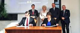 Na fotografii widzimy stolik, przy którym starosta Elżbieta Sadowska oraz Wojewoda Mazowiecki podpisują umowy. w tle stoją parlamentarzyści i widać ekran na którym wyświetlana jest informacja dotycząca Rządowego Funduszu Rozwoju Dróg.