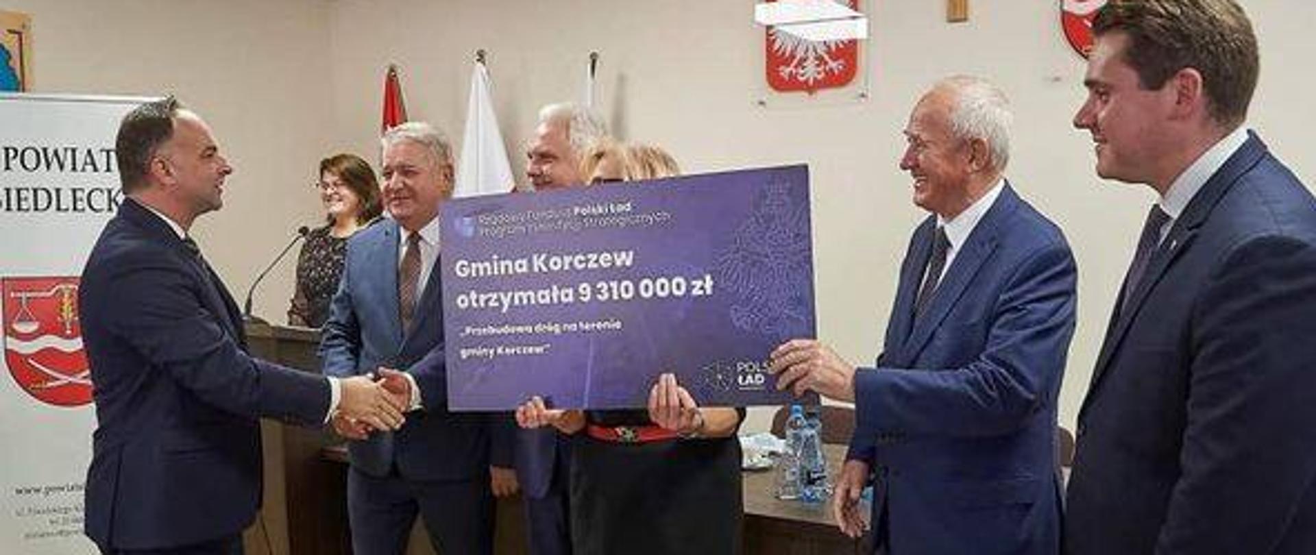 grupa osób. W tle na ścianie godło Polski, krzyż, herb powiatu siedleckiego.Jedna z osób trzyma czek z napisem: Gmina Korczew otrzymała 9 310 000 zł. Przebudowa dróg na terenie gminy Korczew