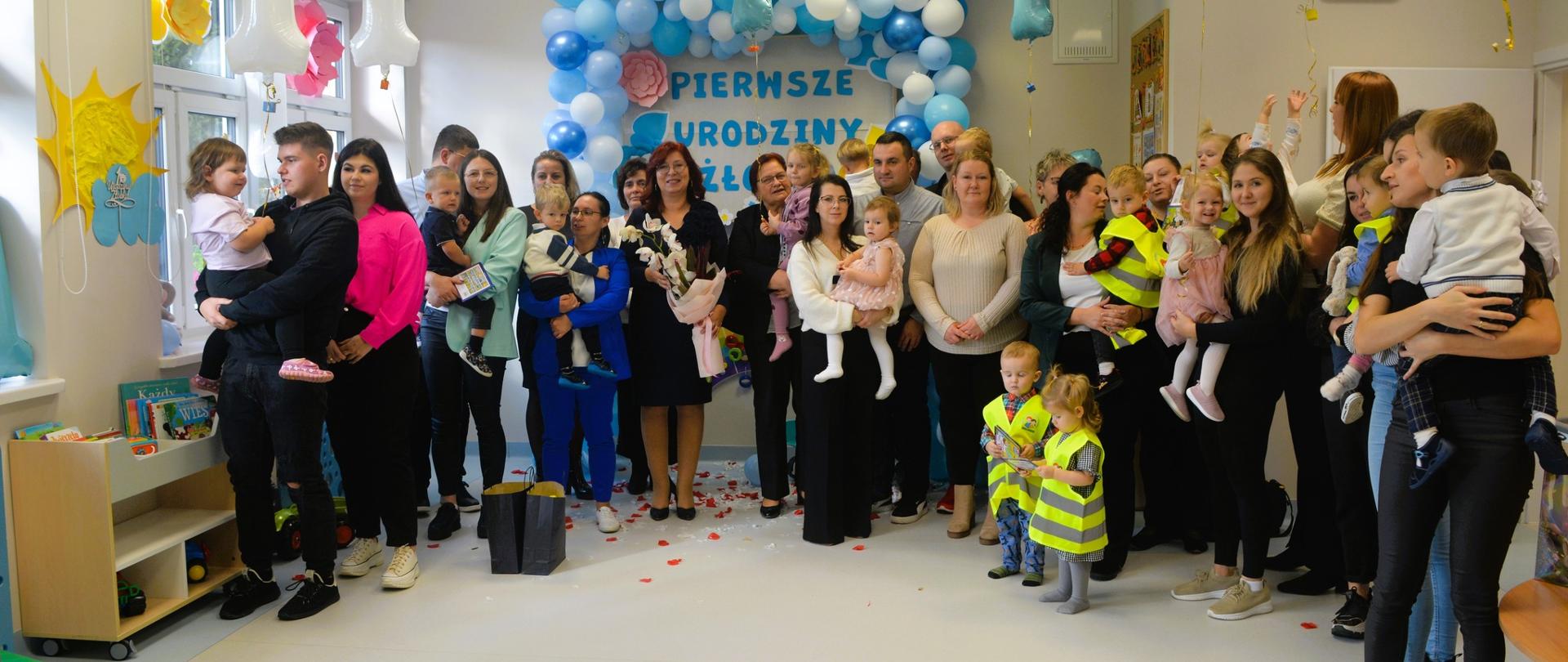 Grupa dorosłych z małymi dziećmi podczas uroczystości. W tle dekoracja z balonów i napis „Pierwsze urodziny żłobka”.