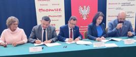Strony umowy siedzą przy nakrytym na zielono stole i podpisują umowy.