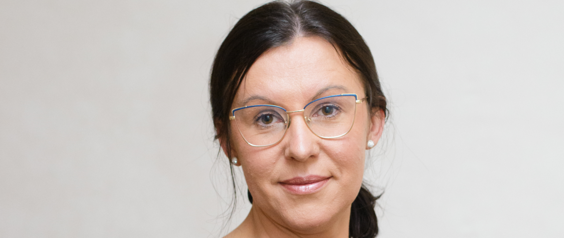 Kobieta w okularach, długie ciemne włosy związane do tyłu, łagodna twarz z lekkim uśmiechem. na uszach białe kolczyki perełki. Ubrana w ciemną bluzkę