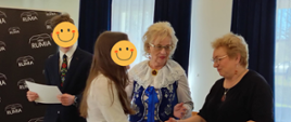 Po prawej stronie widać chłopca i dziewczynkę, mają twarze zasłonięte uśmiechniętymi minkami. Dziewczynka podaje rękę pani stojącej po prawej stronie. Za dziewczynką widać panią ubraną w granatowy kaszubski strój. Panie trzymają w dłoniach papierowe torby z napisem Rumia. Po lewej stronie w tle widać czarne tło z białymi napisami.