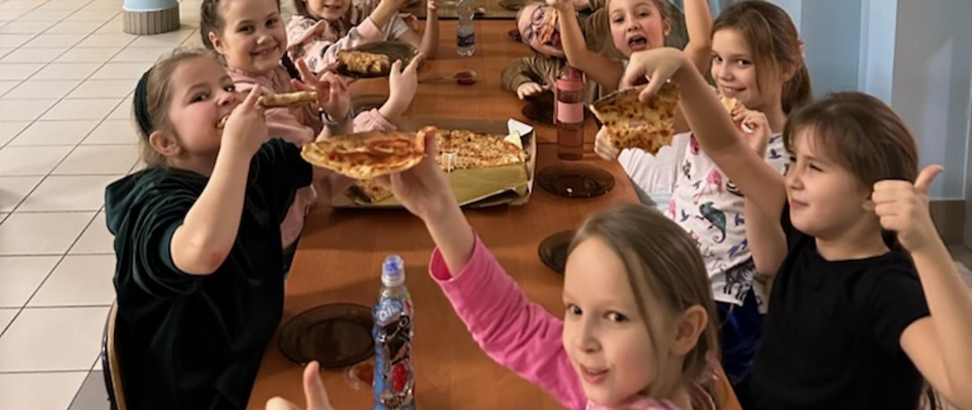 Na zdjęciu są dzieci, które jedzą pizzę. Na pierwszym planie widać dziewczynkę w różowej bluzie o jasnych włosach, dalej siedzą dwie dziewczynki w czarnych koszulkach.
