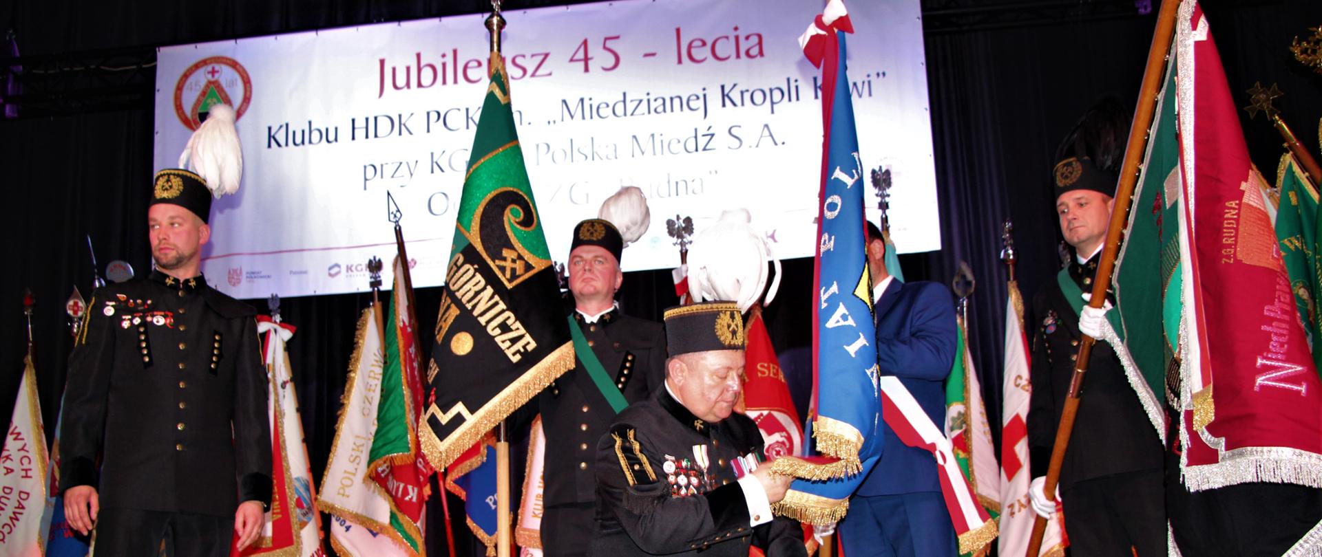 Na scenie stoją ludzie na baczność obok sztandarów. Jeden Pan klęka i trzyma w ręce kawałek sztandaru powiatu polkowickiego.